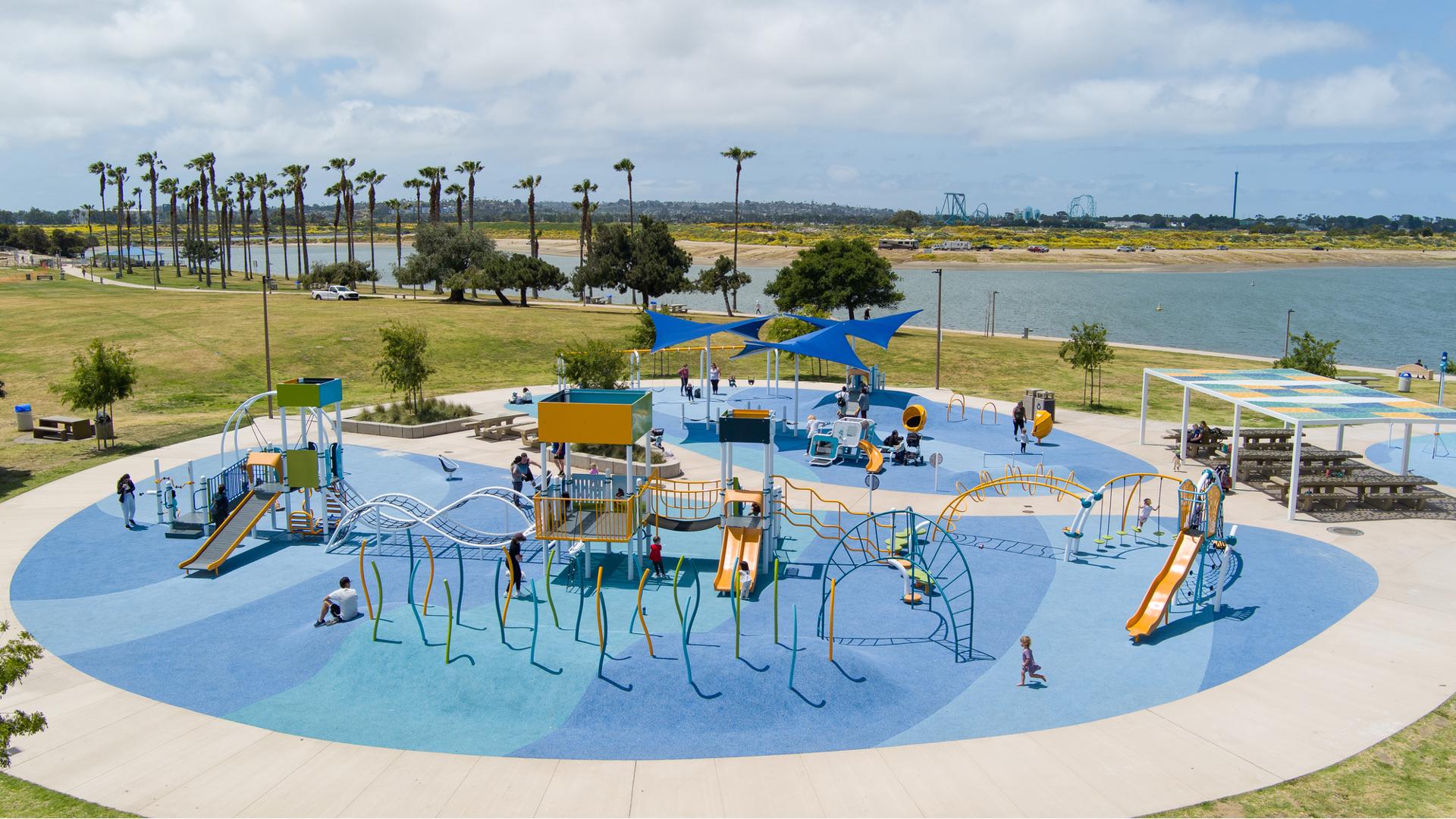 Full elevated view of a large circular play area with multiple play structures and stand alone activities all set next to a large body of water in the background.