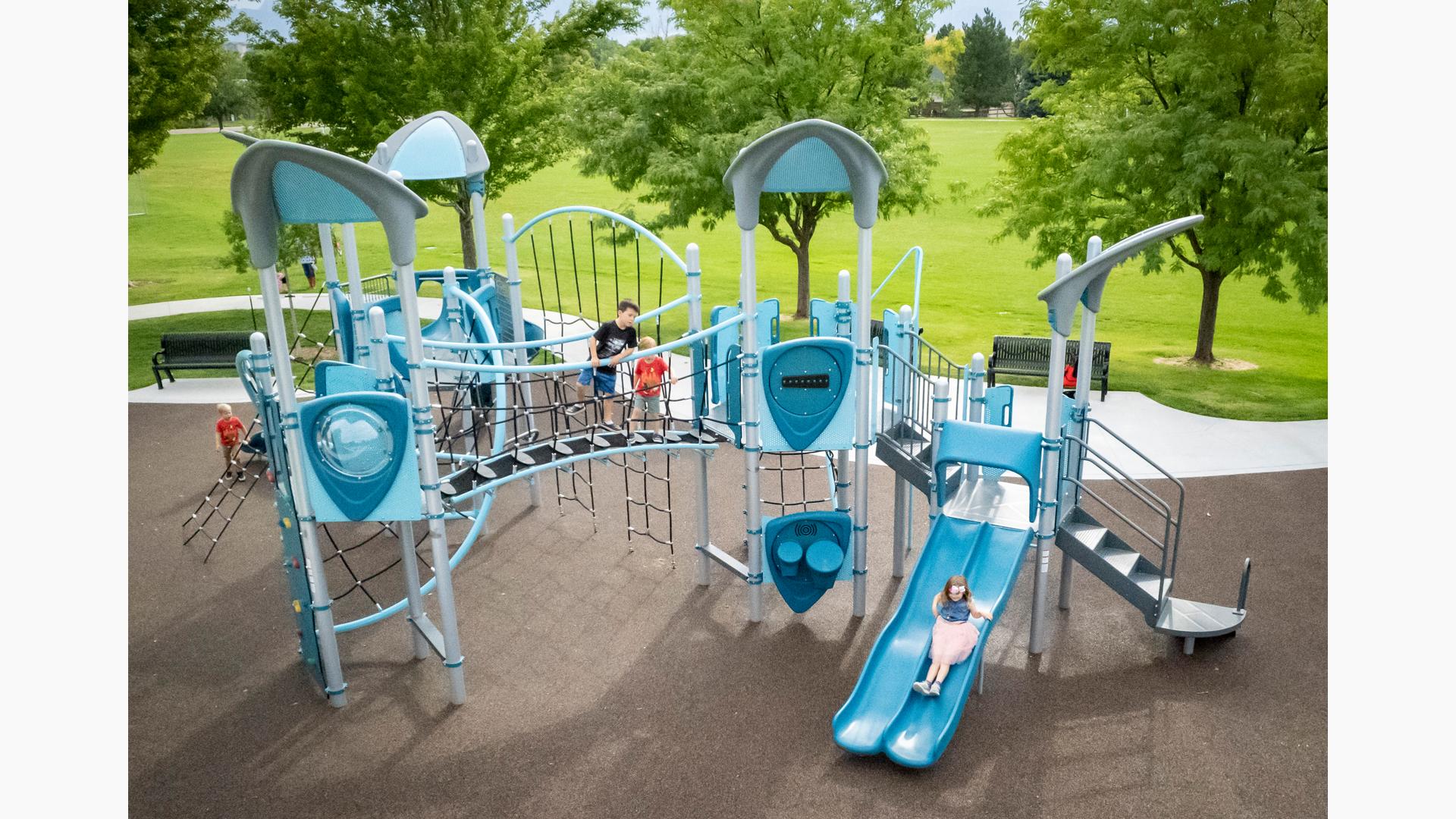 Children play on a park playground colored with different shades of blues and greys with multiple rope climbers, slides, and play panels.