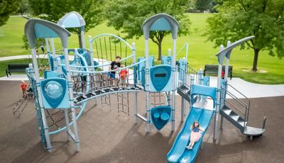 Children play on a park playground colored with different shades of blues and greys with multiple rope climbers, slides, and play panels.