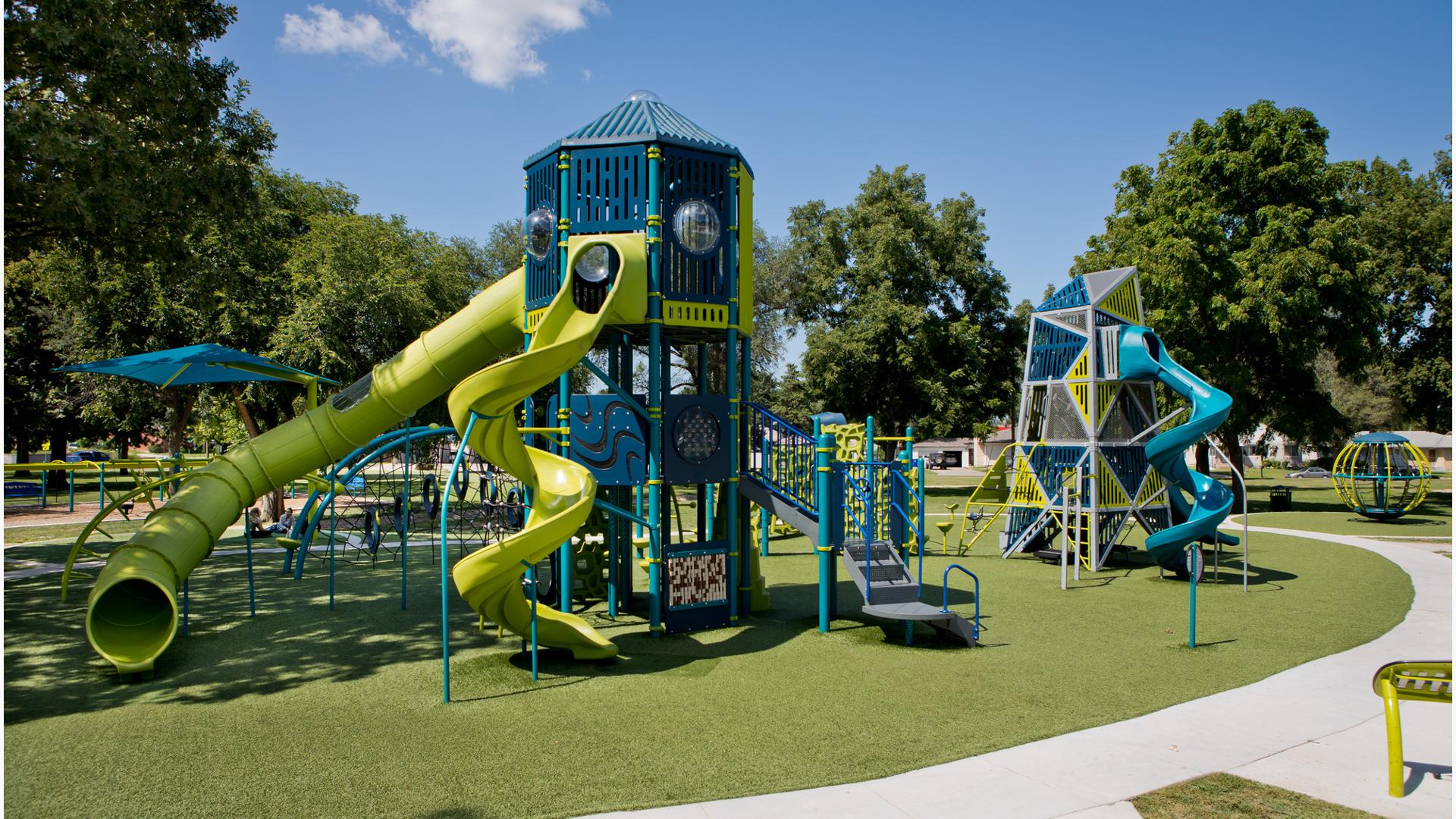 The sun shines on an empty playground while a small group of people sit under a tree and talk amongst themselves.