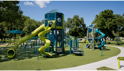 The sun shines on an empty playground while a small group of people sit under a tree and talk amongst themselves.