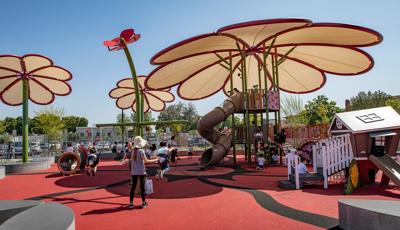 Families play on a playground tower with a large flower shaped shade. Smaller play structures surround the large play tower along with two more large flower shaped shade posts.