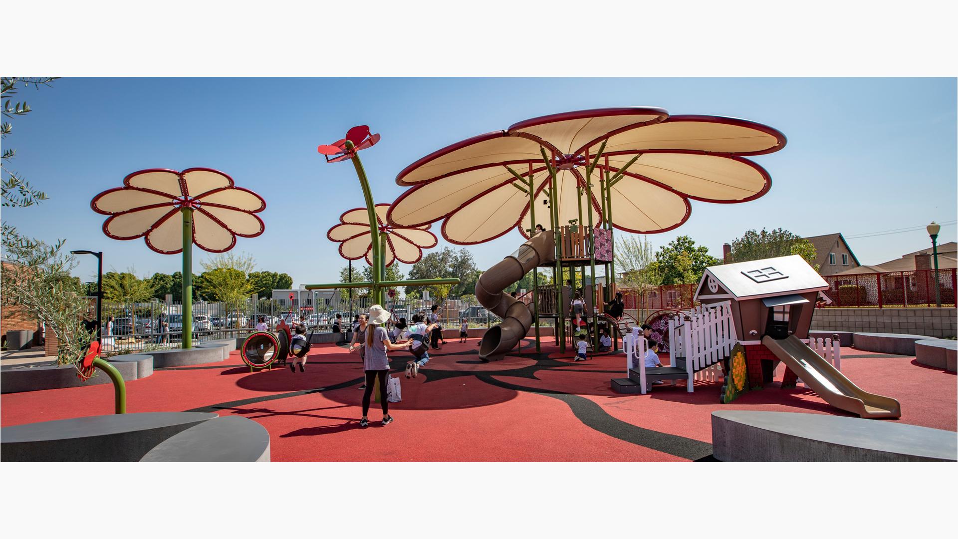 Families play on a playground tower with a large flower shaped shade. Smaller play structures surround the large play tower along with two more large flower shaped shade posts.