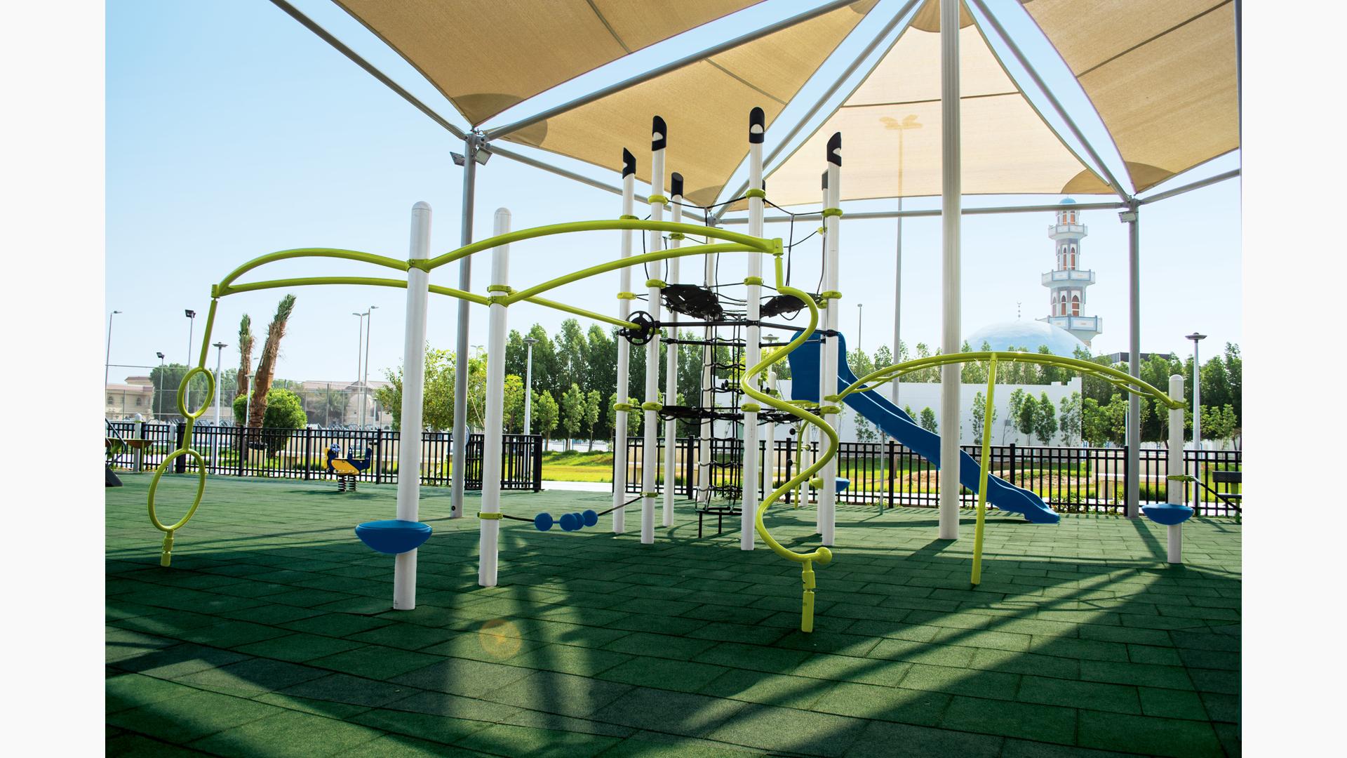 A fenced in play area with a single play structure with additional connected tight rope bridge, monkey bars, and spinners all underneath a large shade system overhead.