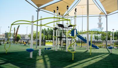 A fenced in play area with a single play structure with additional connected tight rope bridge, monkey bars, and spinners all underneath a large shade system overhead.