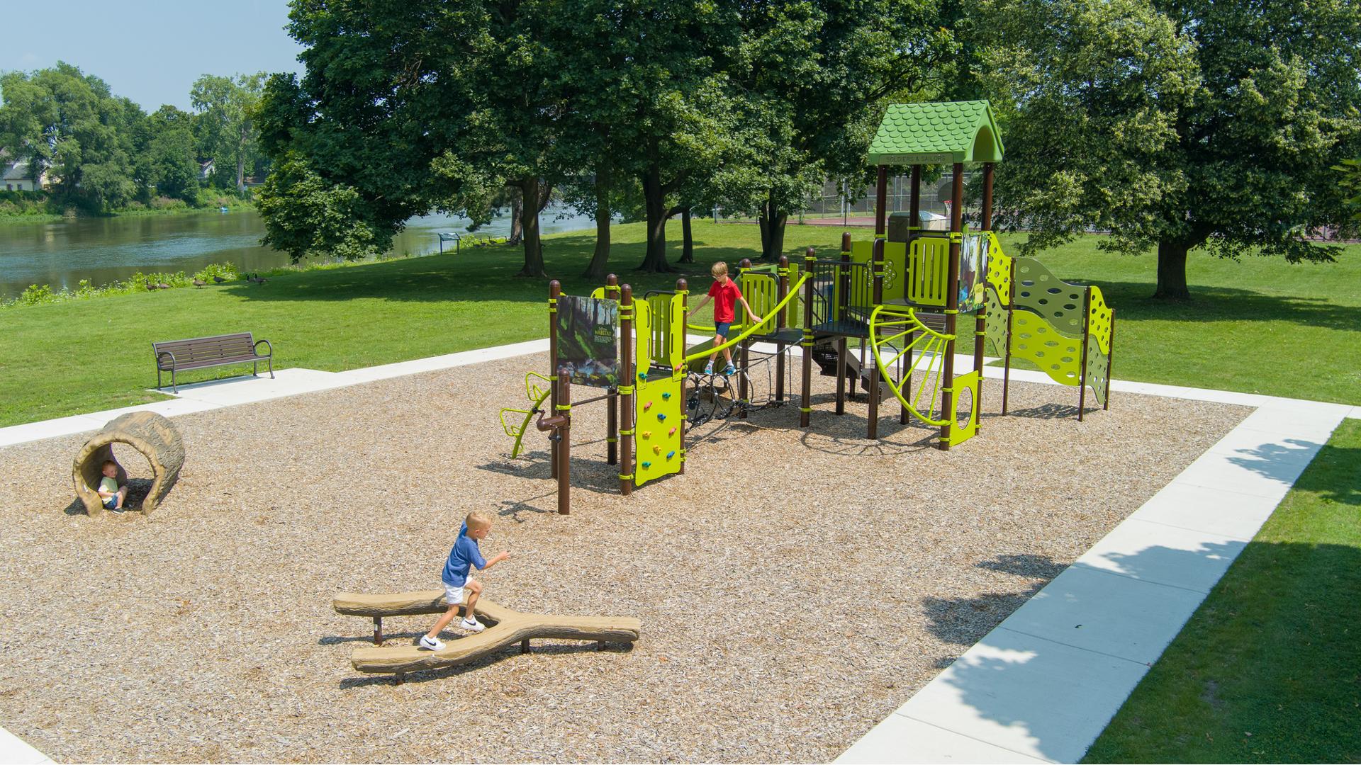 A child plays on a tuning fork shaped log balance beam while other children play on a play structure in the background at a community park play area.