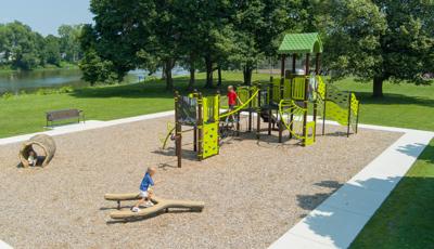 A child plays on a tuning fork shaped log balance beam while other children play on a play structure in the background at a community park play area.