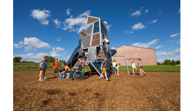 A boy sits on the bottom step of the Alpha® Tower talking to 3 girls. A boy in orange shorts walks in front of  Alpha® Tower. Other children in the background climb in and on this skyscraper-like structure.