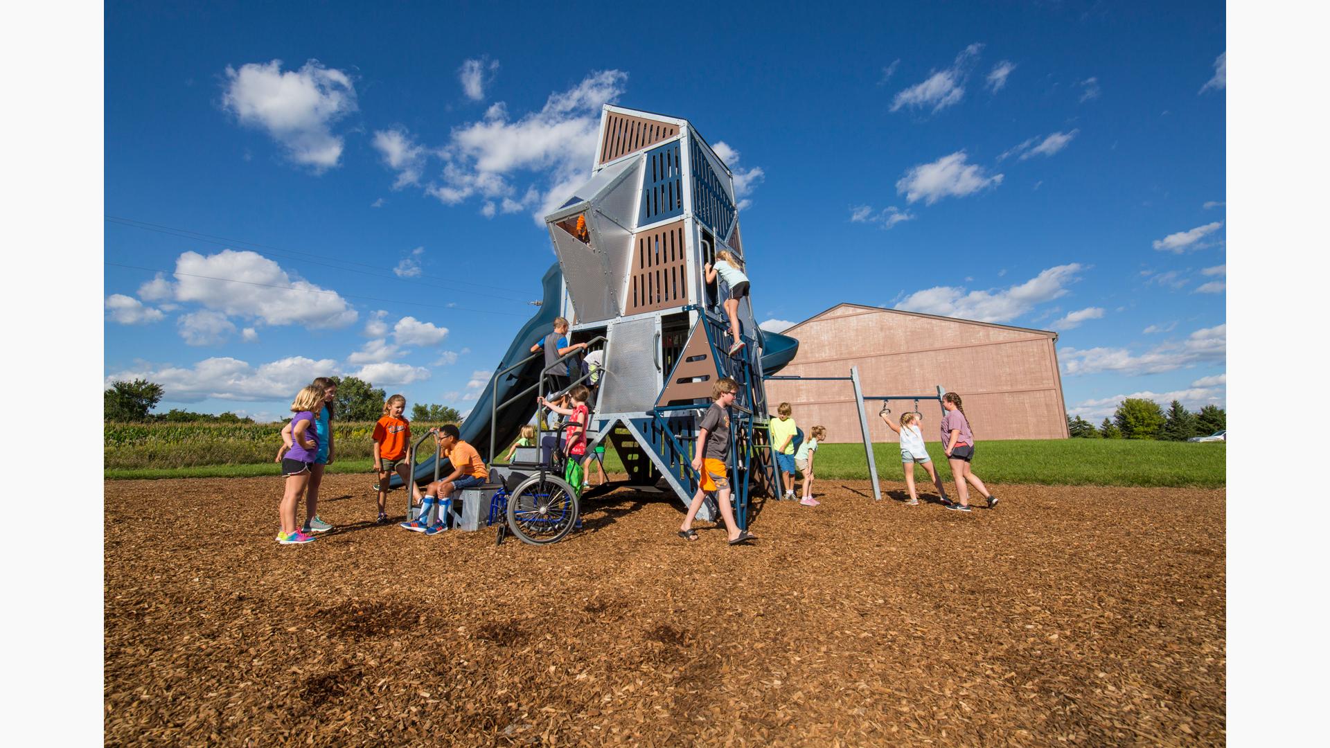 A boy sits on the bottom step of the Alpha® Tower talking to 3 girls. A boy in orange shorts walks in front of  Alpha® Tower. Other children in the background climb in and on this skyscraper-like structure.