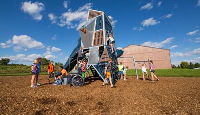 A boy sits on the bottom step of the Alpha® Tower talking to 3 girls. A boy in orange shorts walks in front of  Alpha® Tower. Other children in the background climb in and on this skyscraper-like structure.