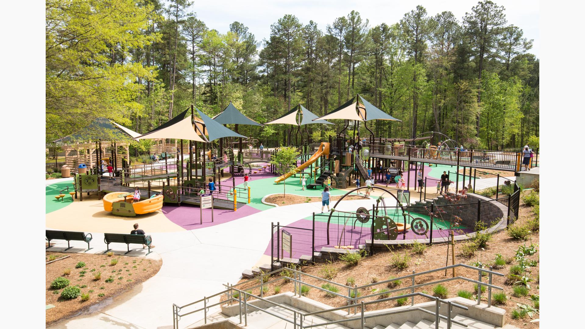 Filled with bridges and ramps, the playground also has tan and green multi-panel shade structures. This is a large playground with orange slides and surrounded by tall green trees.