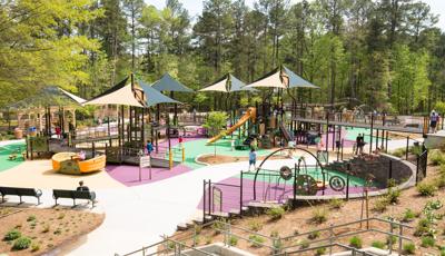 Filled with bridges and ramps, the playground also has tan and green multi-panel shade structures. This is a large playground with orange slides and surrounded by tall green trees.
