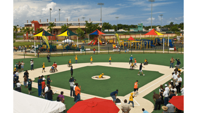 Parents and kids on baseball field playing a lively game. Large, colorful playground with shade and many ramps in background. Lots of slides and equipment as kids play all around.