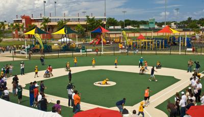 Parents and kids on baseball field playing a lively game. Large, colorful playground with shade and many ramps in background. Lots of slides and equipment as kids play all around.