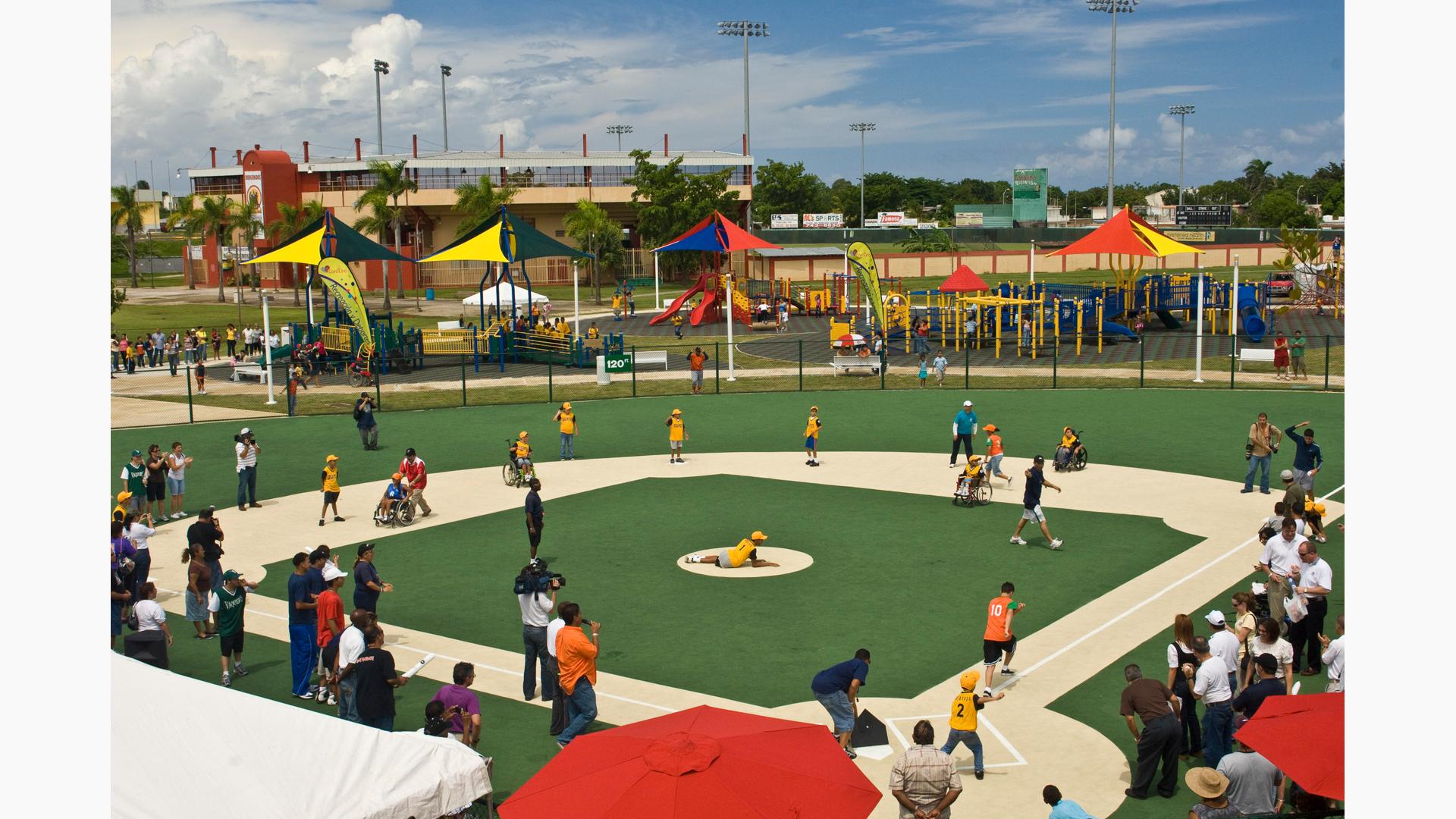Parents and kids on baseball field playing a lively game. Large, colorful playground with shade and many ramps in background. Lots of slides and equipment as kids play all around.