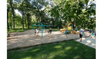 Families playing in Wildwood Park. Mothers pushing their toddlers on swings while a dad watches his child play on a Smart Play play structure.