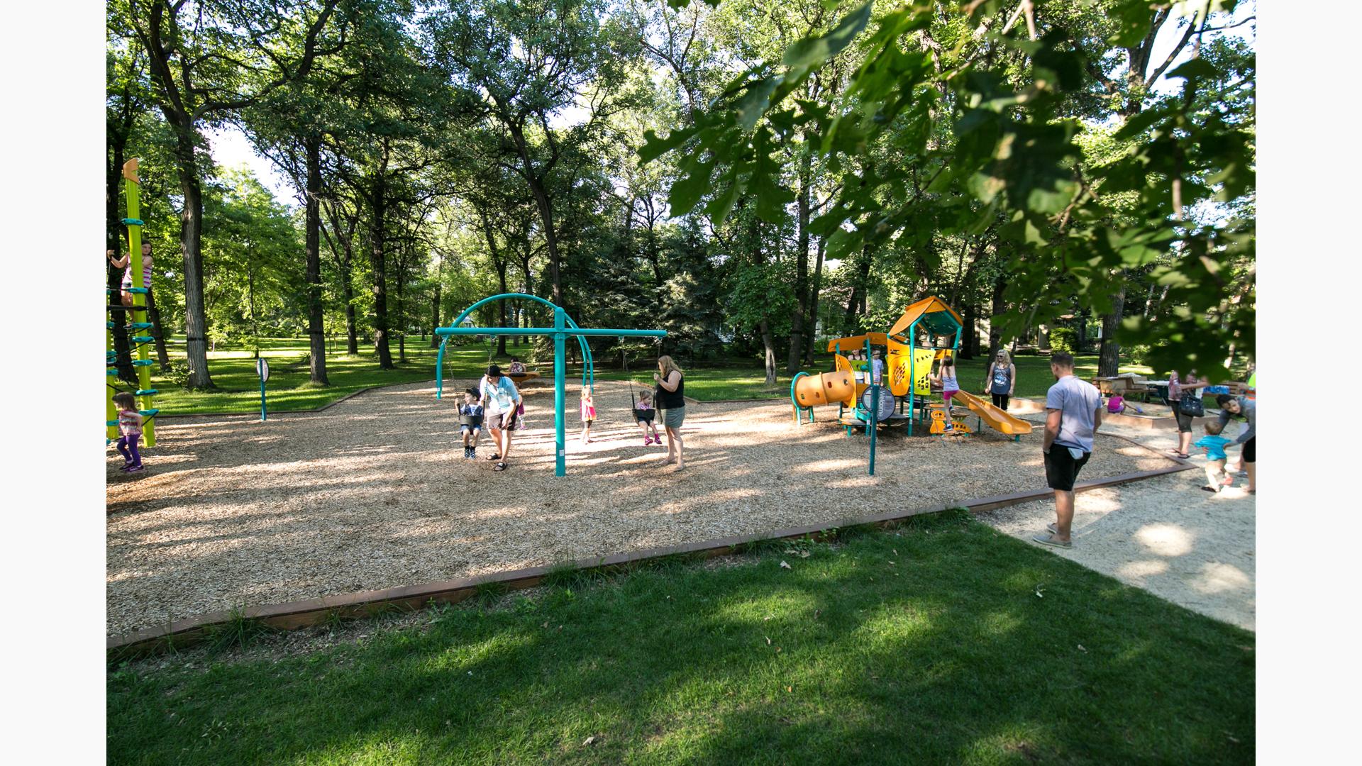 Families playing in Wildwood Park. Mothers pushing their toddlers on swings while a dad watches his child play on a Smart Play play structure.