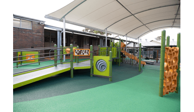 A play area with green safety surfacing with a play structure made up of accessible bridges and ramps leading to play panels, slides, and climbers. The play area is completely covered by a large arched shade next to a brick building. 