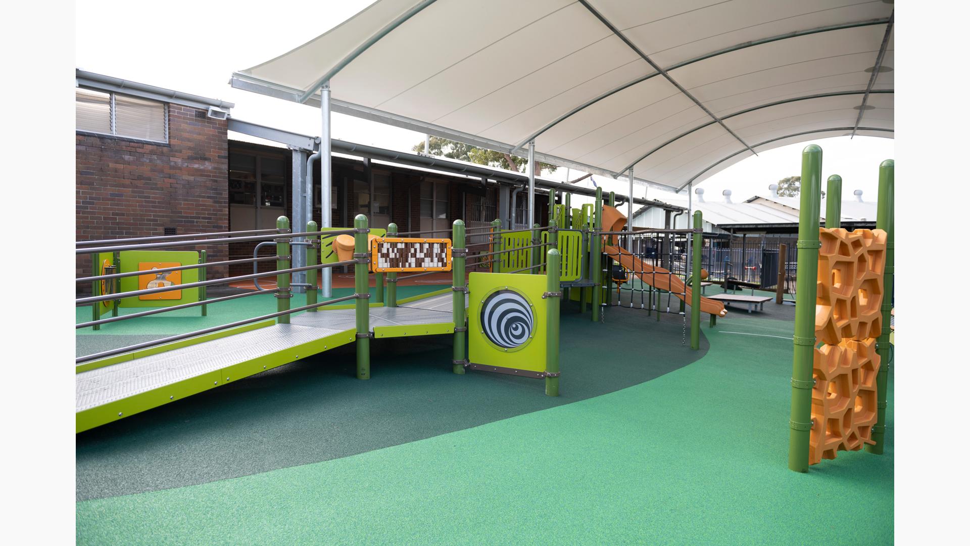 A play area with green safety surfacing with a play structure made up of accessible bridges and ramps leading to play panels, slides, and climbers. The play area is completely covered by a large arched shade next to a brick building. 