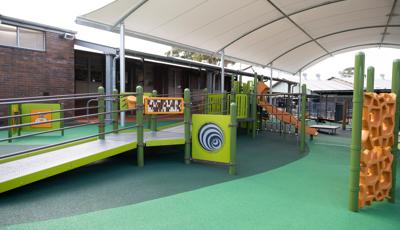 A play area with green safety surfacing with a play structure made up of accessible bridges and ramps leading to play panels, slides, and climbers. The play area is completely covered by a large arched shade next to a brick building. 