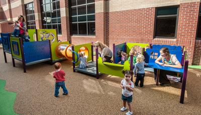 Toddlers play with their teachers at multiple play panels making up a play structure next to a brick building. 