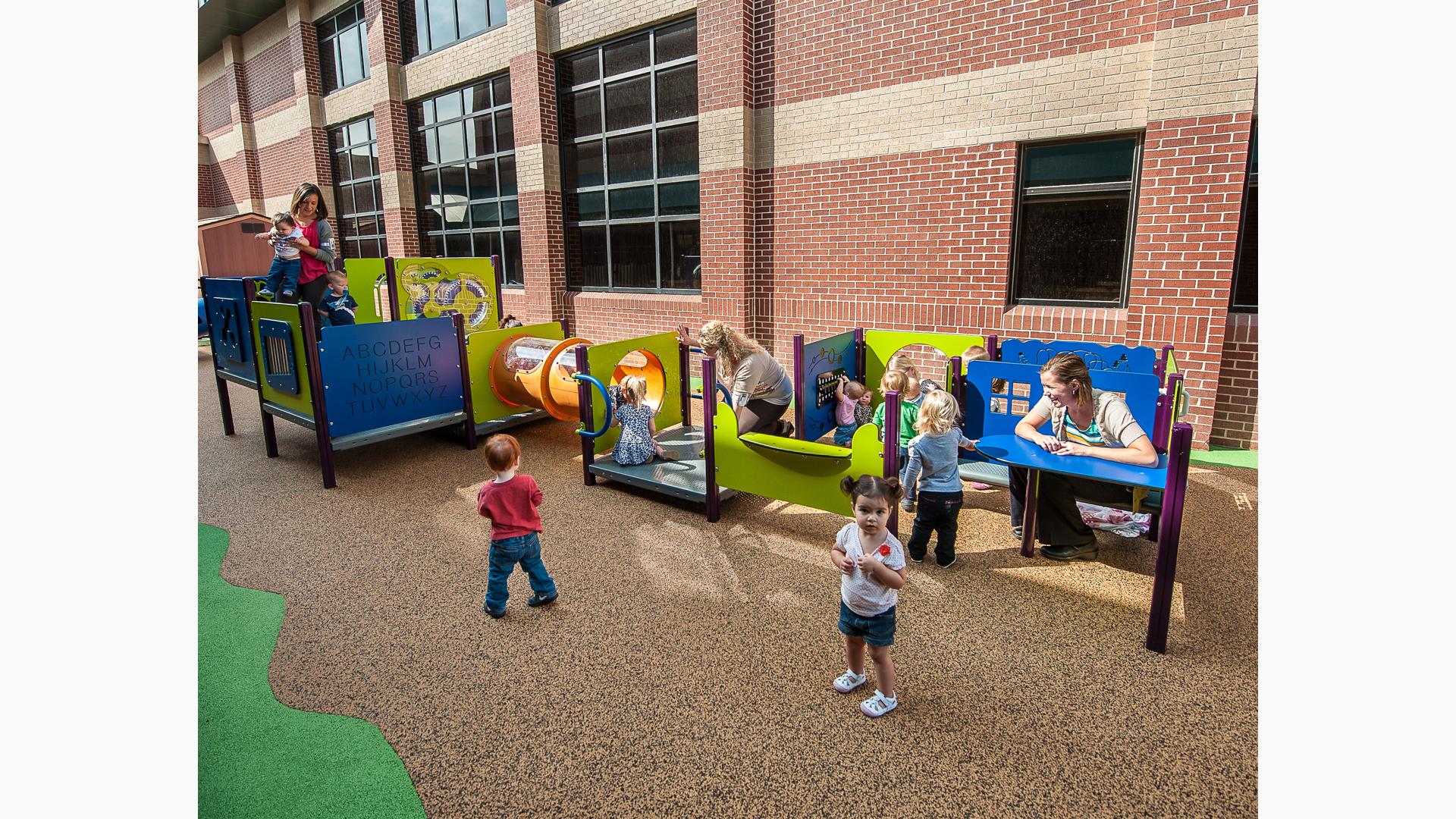 Toddlers play with their teachers at multiple play panels making up a play structure next to a brick building. 