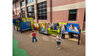 Toddlers play with their teachers at multiple play panels making up a play structure next to a brick building. 