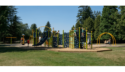 Lots of kids playing on a playground with navy blue geometric climbers and slides. Kids swinging in the background in amongst tall green pine trees. 