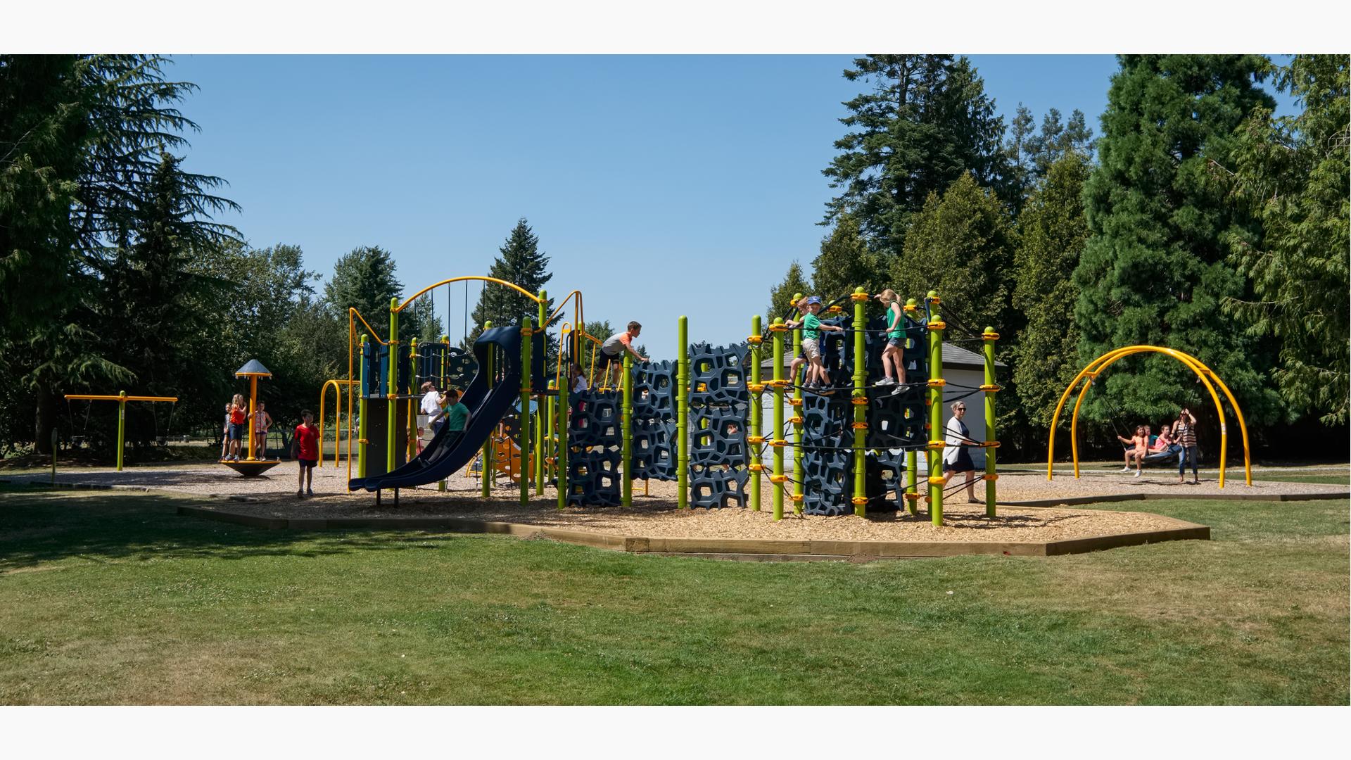 Lots of kids playing on a playground with navy blue geometric climbers and slides. Kids swinging in the background in amongst tall green pine trees. 