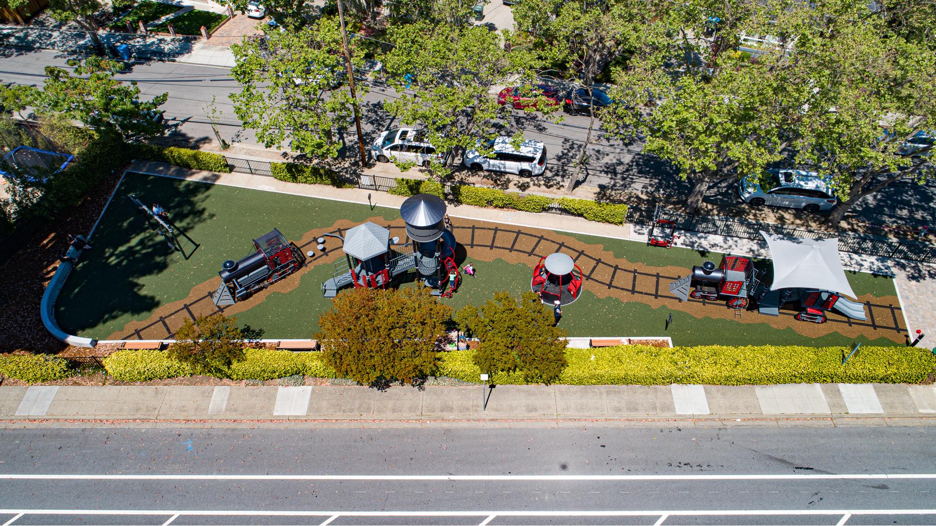 Full bird's eye view of a long and narrow play area with train designed play structures on train track designed playground surfacing. 