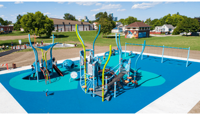 Full elevated view of a park playground with underwater themed play structures, children play at a splash pad just beyond the playground area.
