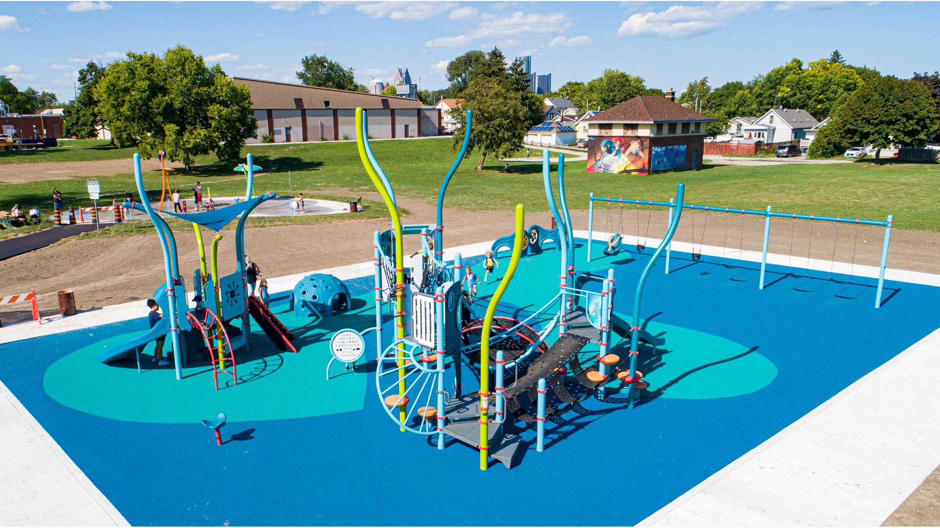 Full elevated view of a park playground with underwater themed play structures, children play at a splash pad just beyond the playground area.