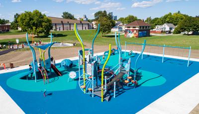 Full elevated view of a park playground with underwater themed play structures, children play at a splash pad just beyond the playground area.