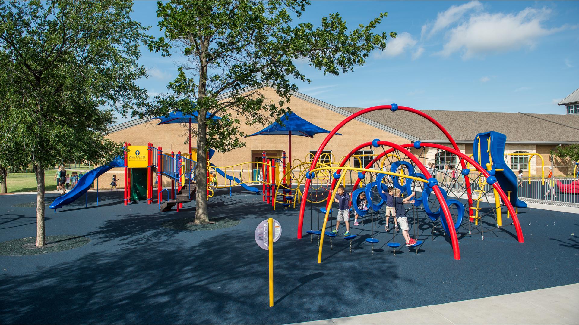 Three children play on arched posts of a play structure with rope climbers and an additional larger play structure in the background closest to a school brick building.