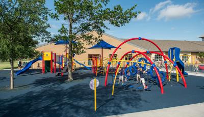 Three children play on arched posts of a play structure with rope climbers and an additional larger play structure in the background closest to a school brick building.