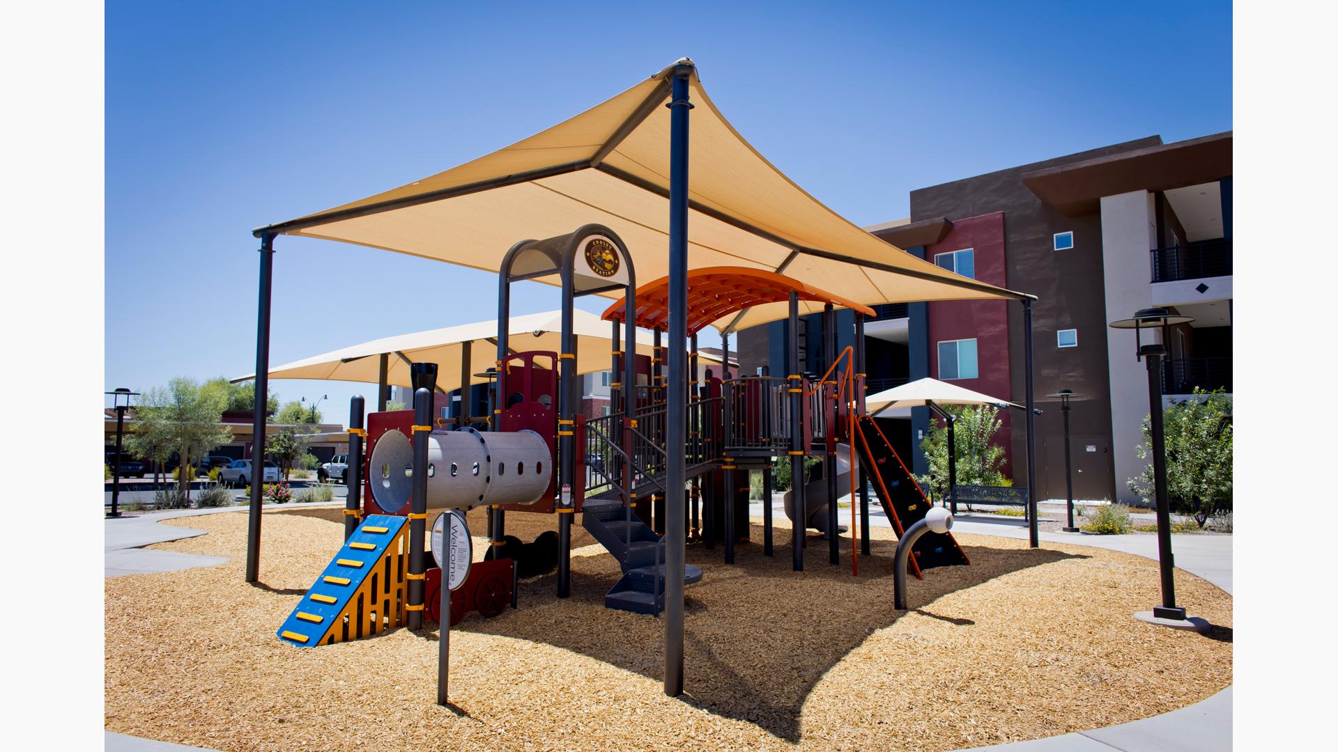 A play structure designed like an imaginative train with a large rectangular shade overhead and woodchip ground surfacing. The playground is set next to a residential building.