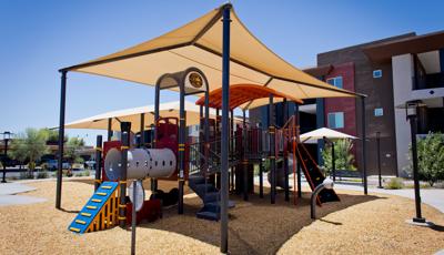 A play structure designed like an imaginative train with a large rectangular shade overhead and woodchip ground surfacing. The playground is set next to a residential building.