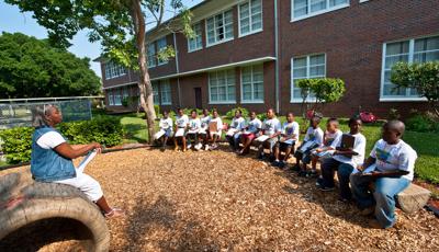 A woman sitting on a log designed crawl tunnel speaks to a class of children with clipboards sitting across from her on split log benches in an outdoor courtyard. 