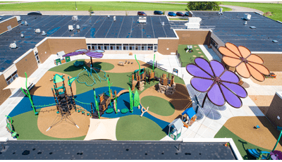 Elevated view of a nature themed central playground surrounded by a school building equipped with two large flower shaped shade systems.