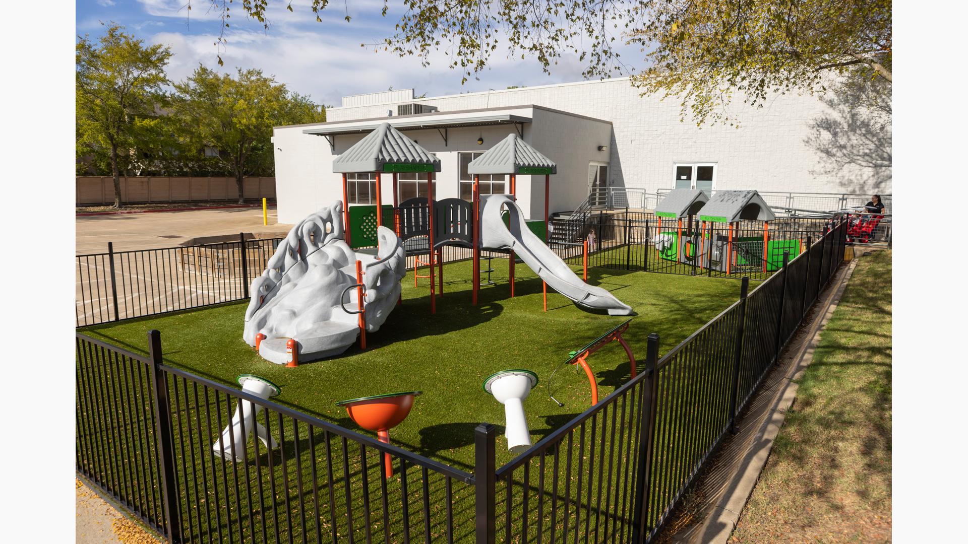 A black iron fence surrounds a play area with play structure, outdoor musical instruments and play panels for young children outside a white brick building. 