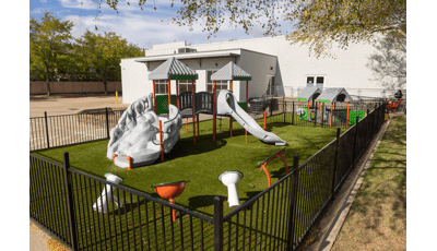 A black iron fence surrounds a play area with play structure, outdoor musical instruments and play panels for young children outside a white brick building. 
