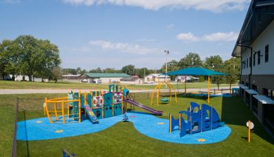 A play area sits on the back side of a building surrounded by a chain link fence. In the foreground a play structure designed like an imaginative boat with an additional play structure shaped like a fish to the right. In the background a square shade stands over a few picnic tables.