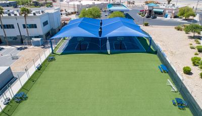 Elevated view of a fenced in artificially grassed play area with two side by side basketball courts covered by two large blue rectangular shade roofs.