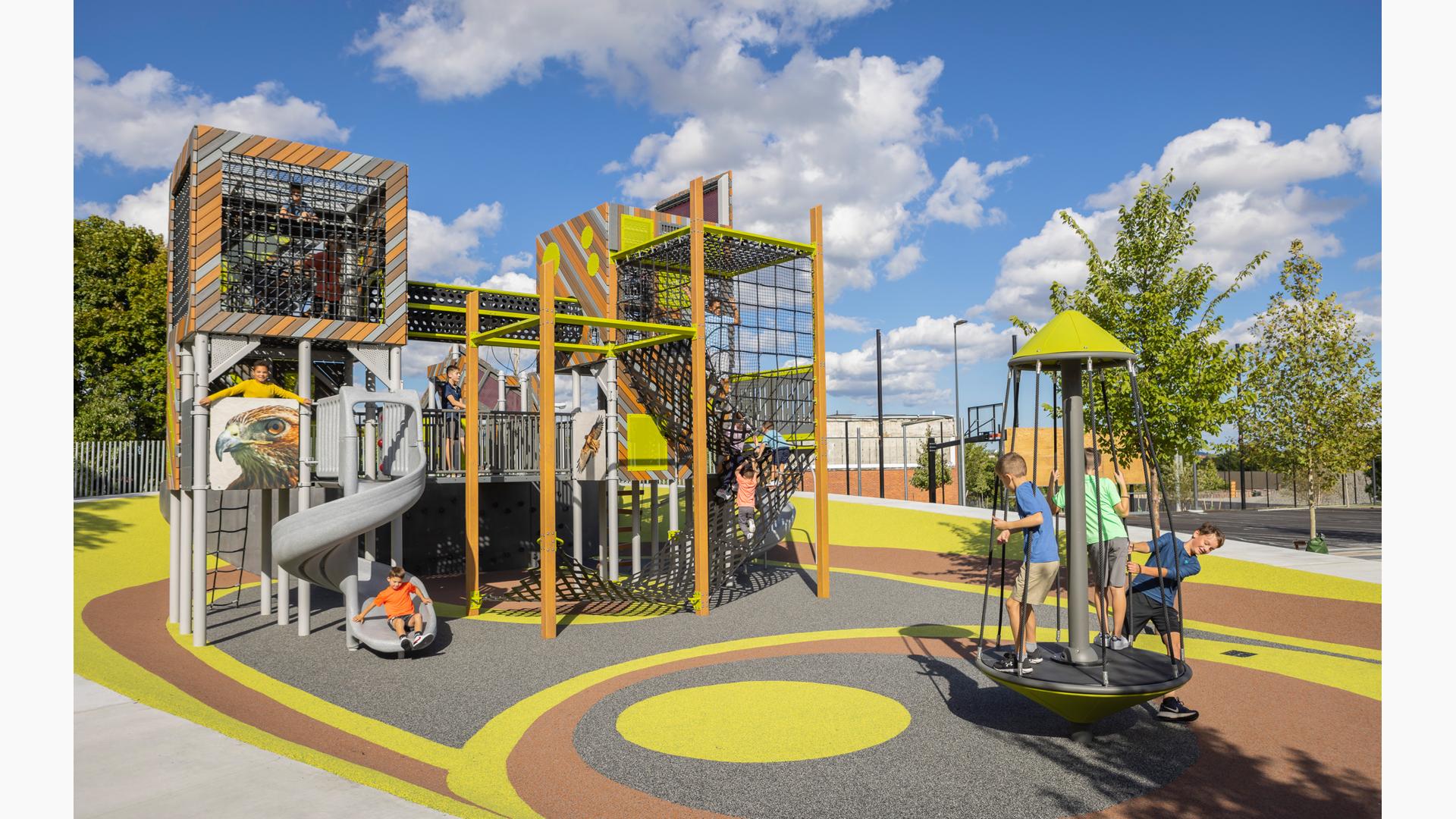 Children play on a multi rider standing spinner next to a unique modern designed play structure made with recycled lumber panels and square enclosed towers.