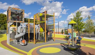 Children play on a multi rider standing spinner next to a unique modern designed play structure made with recycled lumber panels and square enclosed towers.