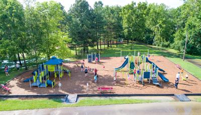 Elevated view of a large park play area with two separate play structures for different age children with additional swing set and climbing wall behind them. The park is filled with playing families.