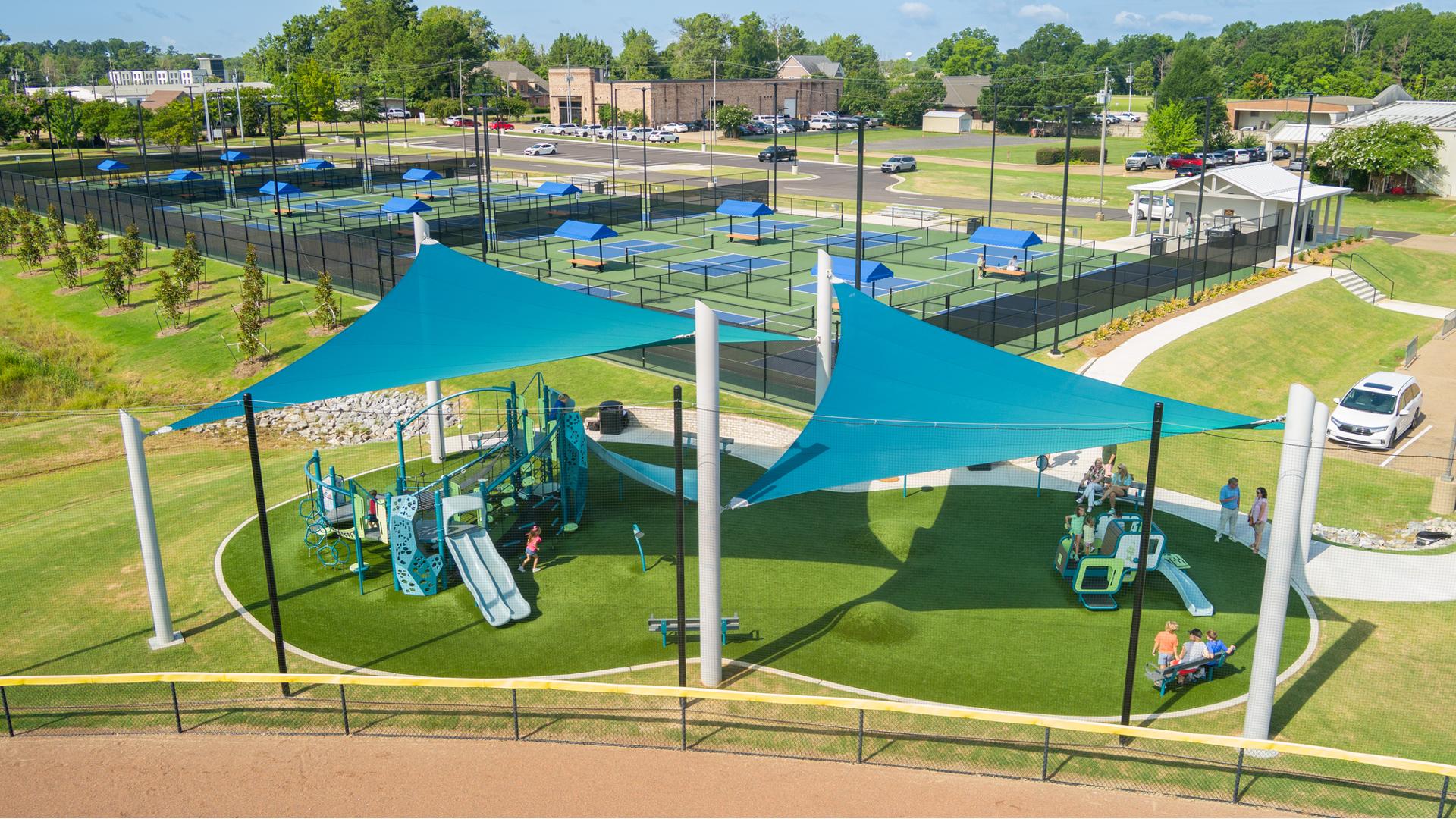 Elevated view of a playground with two large shade sails overhead all next to pickle ball courts.