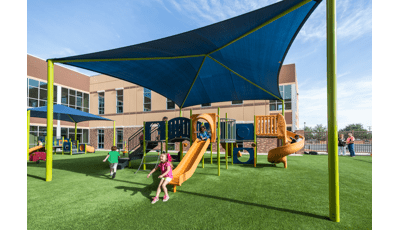 Girl in pink jumps off slide while other kids run around to get back in line to ride it again. A large square blue shade covers the play structure. 