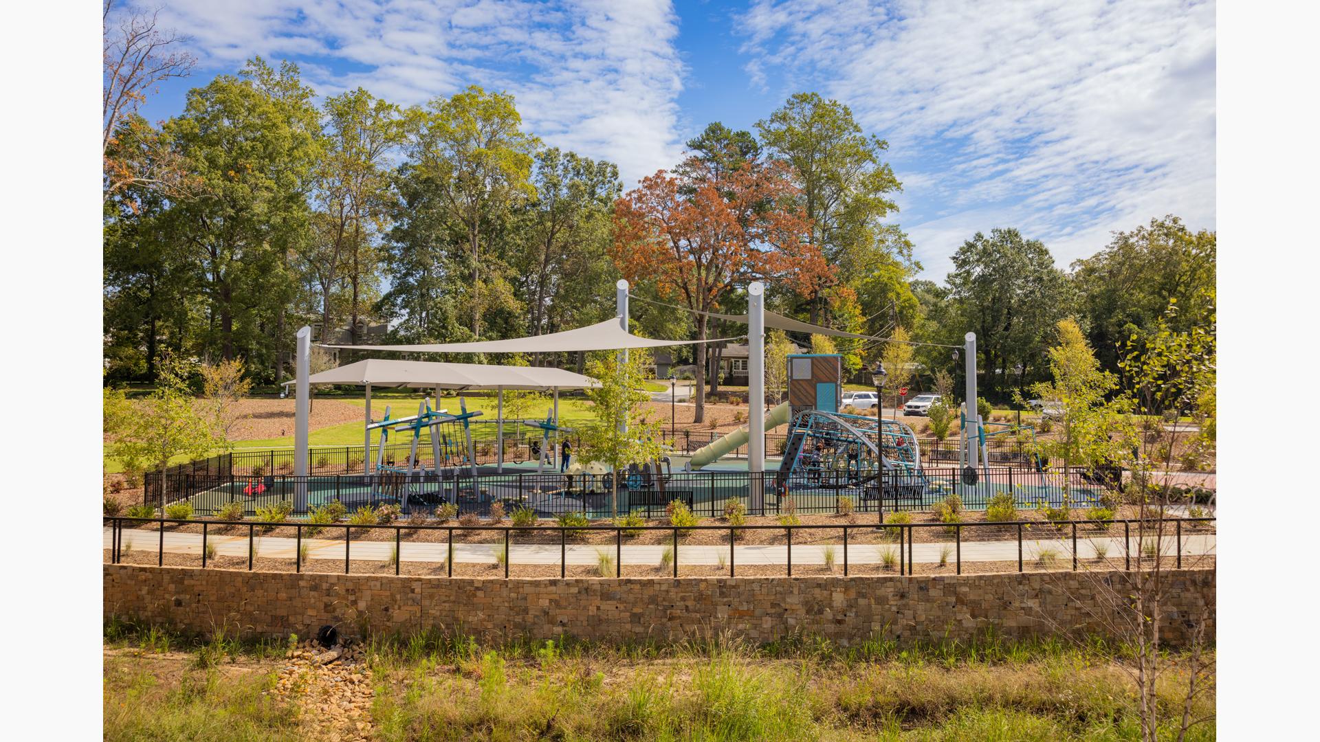 A playground with large grey shade sails overhead surrounded by landscaping and a black fence.