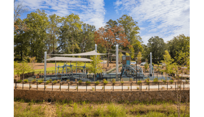 A playground with large grey shade sails overhead surrounded by landscaping and a black fence.